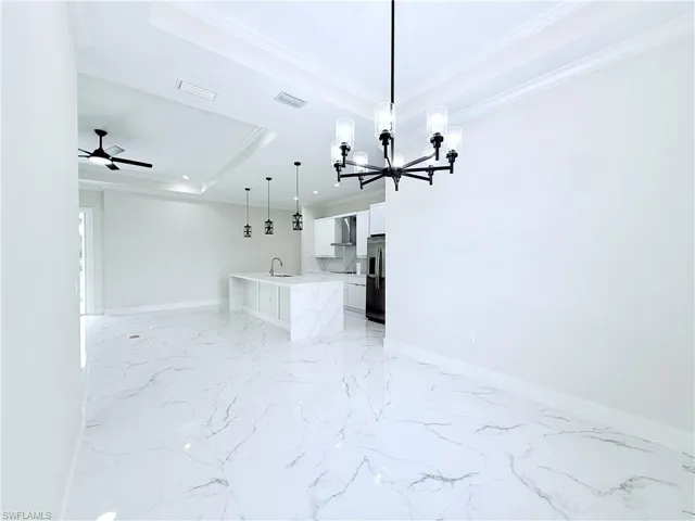 Unfurnished living room featuring a sink, a chandelier, a raised ceiling, light marble finish floors, and crown molding