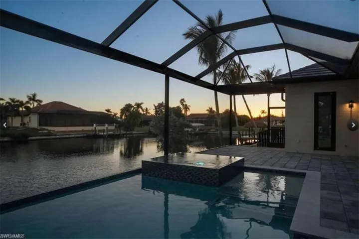 Pool at dusk with glass enclosure, an in ground hot tub, a water view, and a patio