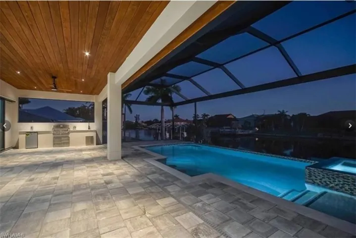 Pool at night featuring exterior kitchen, ceiling fan, a patio, and glass enclosure