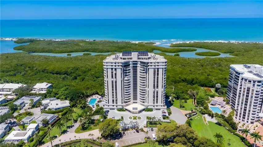 High-rise building with a curved façade and multiple balconies, featuring a circular drive, lush green landscaping, and an outdoor swimming pool, all against a backdrop of a clear blue ocean and dense coastal foliage