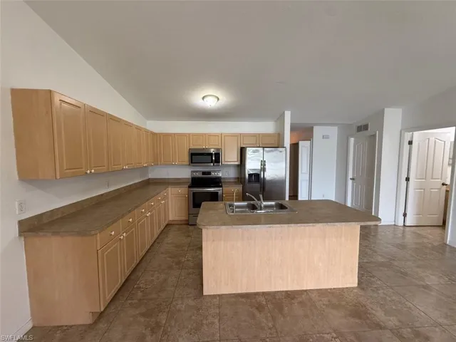 Kitchen with appliances with stainless steel finishes, light brown cabinetry, a center island with sink, and lofted ceiling