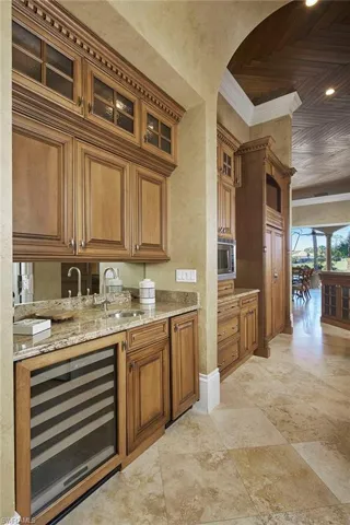 Bar area featuring brown cabinetry, beverage cooler, light stone countertops, glass insert cabinets, and ornamental molding