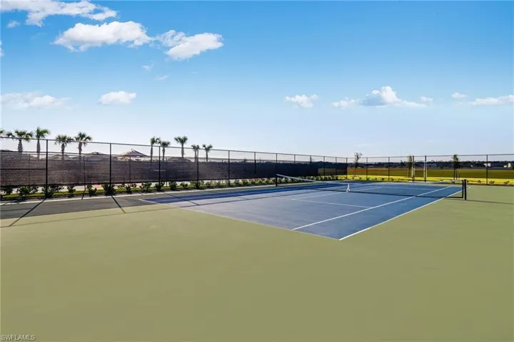 Tennis court featuring a blue playing surface, green surrounding areas, black chain-link fencing, and a privacy screen