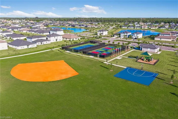 Expansive green space featuring a vibrant orange baseball field, a blue-surfaced basketball court, a dedicated playground area with a shade canopy, and multiple fenced tennis courts