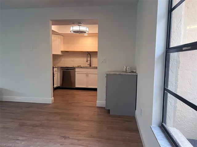 Kitchen with white cabinetry, stainless steel dishwasher, dark wood-style flooring, light stone counters, and backsplash
