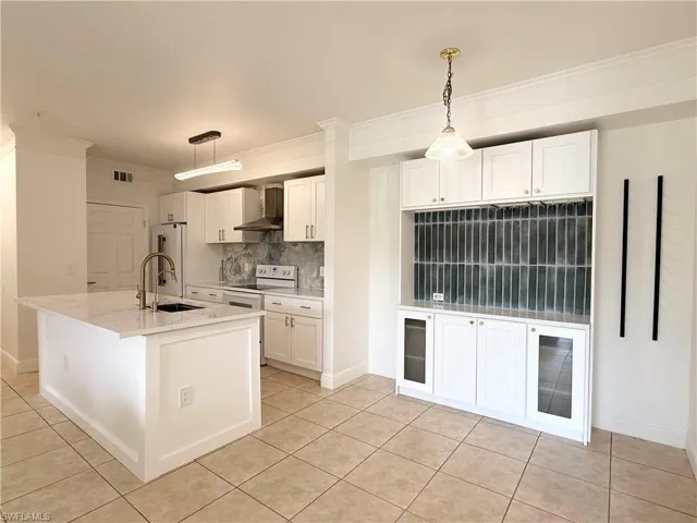 Kitchen featuring a central island with a sink and gold-tone faucet, white cabinetry, integrated range hood, tile flooring, and a built-in display unit with glass-front cabinets and a tiled backsplash
