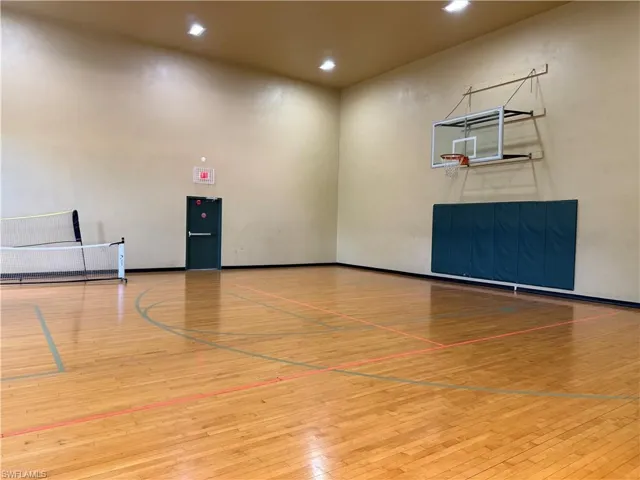 Indoor sports court featuring hardwood flooring with court markings, a wall-mounted basketball hoop with backboard padding, and recessed ceiling lighting