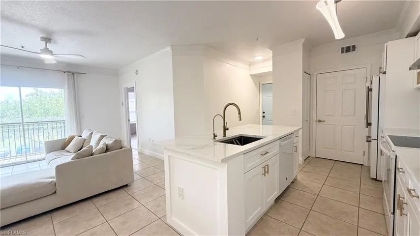 Open-concept kitchen and living area featuring a central island with a sink and faucet, white cabinetry with brass hardware, a full suite of white appliances, and tiled flooring