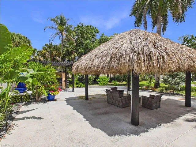 Thatch-roofed structure over a concrete patio featuring tropical landscaping and a black pergola