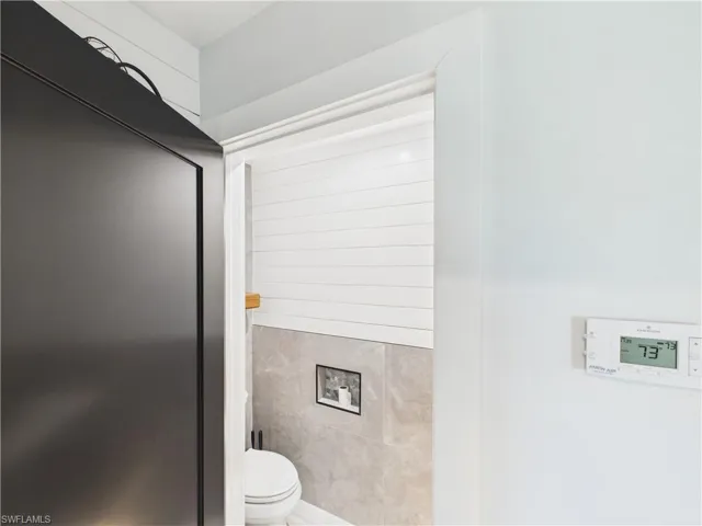 Recessed wall-mounted toilet with a dark-framed flush plate, grey stone-finish wall tile, white shiplap paneling, and a wood shelf