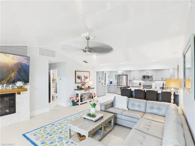 Open-concept living area featuring light-toned flooring, a fireplace with a wood mantle, a ceiling fan, and a kitchen with light grey cabinetry, stainless steel appliances, and a breakfast bar