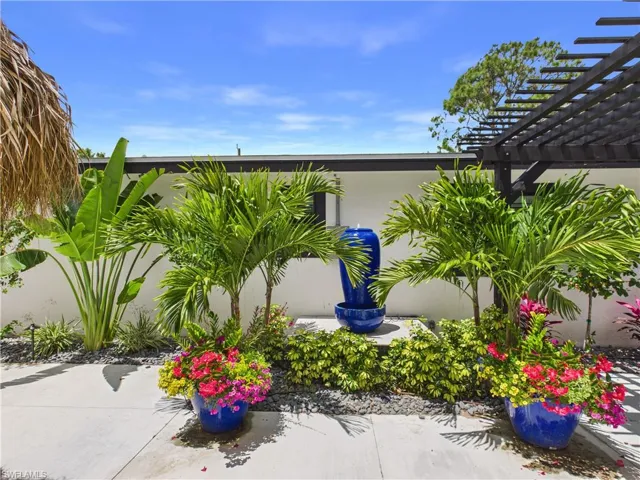 Landscaped courtyard featuring a tiered blue ceramic fountain, a dark-stained wood pergola, lush tropical foliage, and vibrant potted floral arrangements