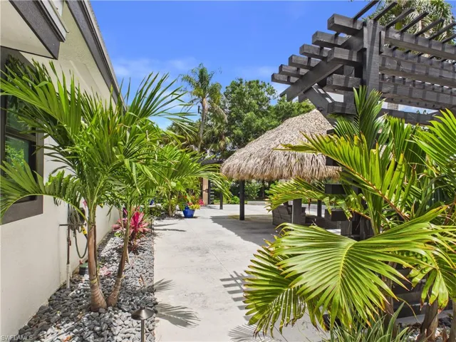 Outdoor space featuring a thatched-roof structure, dark-stained pergola, concrete pathway, and lush tropical landscaping