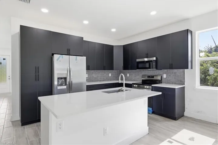 Modern kitchen featuring dark cabinetry, stainless steel appliances, a white island with an integrated sink, a gray subway tile backsplash, and light wood-finish flooring