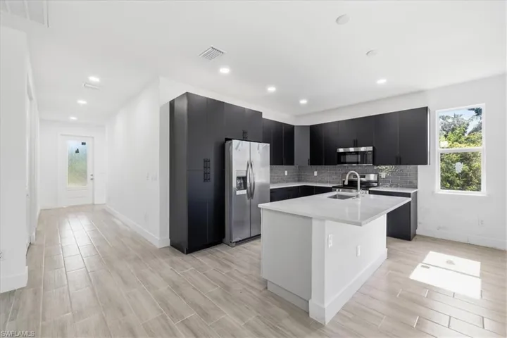 Modern kitchen featuring dark cabinetry with brushed nickel hardware, a central island with a sink, stainless steel appliances, and a gray subway tile backsplash