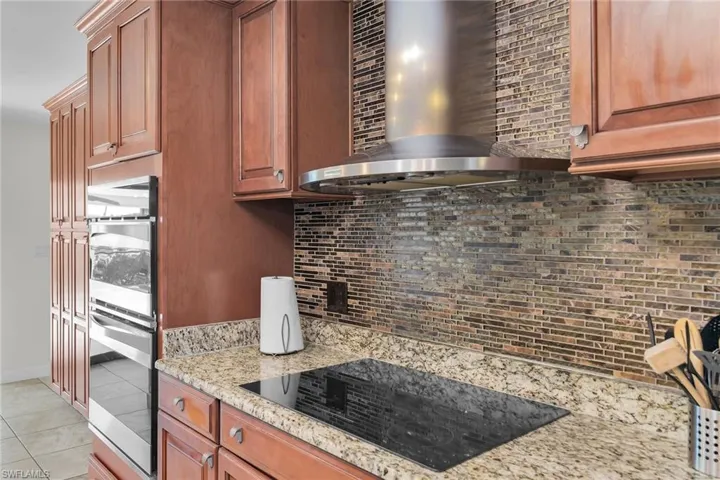 Kitchen with wall chimney exhaust hood, black electric stovetop, light stone countertops, light tile patterned flooring, and double oven