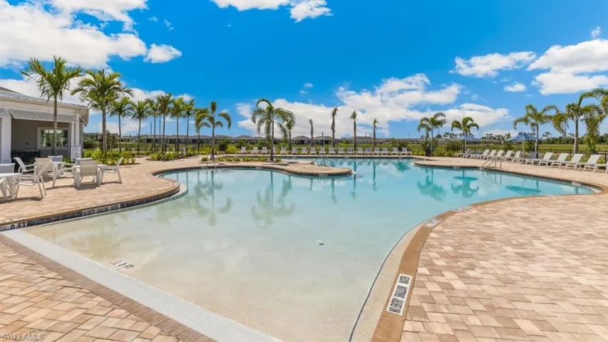 Expansive resort-style swimming pool featuring a shallow entry area, surrounded by a paved deck with chaise lounges and palm trees, with a building visible in the background