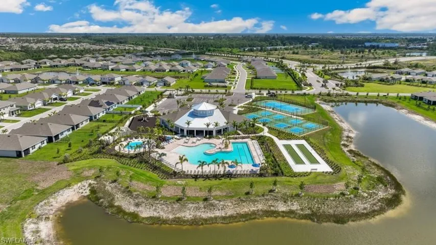 An aerial view showcasing the community's extensive amenities, including a large clubhouse, two swimming pools, tennis courts, and a bocce ball court, all set against a backdrop of residential properties and natural waterways