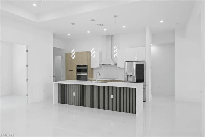 Kitchen featuring modern cabinets, an island with sink, hanging light fixtures, light stone counters, and wall chimney range hood