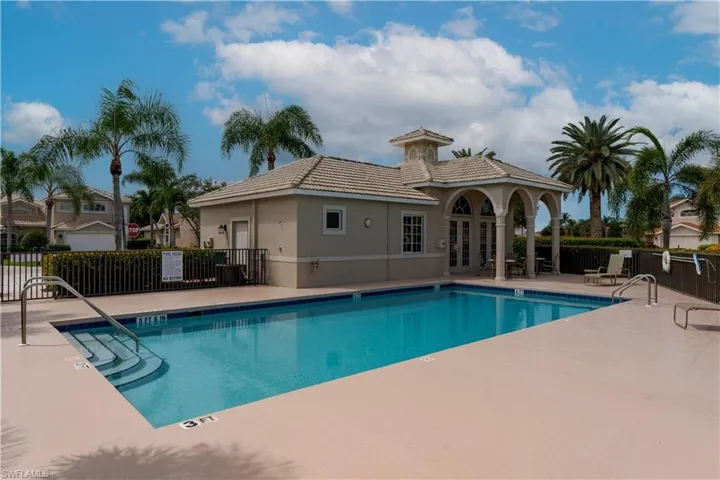 Resort-style pool area featuring a large rectangular pool with integrated steps, a surrounding paved deck, and a clubhouse with a covered patio supported by archways