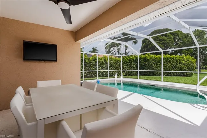 Covered outdoor patio with a ceiling fan, wall-mounted television, and direct access to a screened-in pool area featuring a rectangular swimming pool, white deck, and surrounding manicured lawn with mature hedges