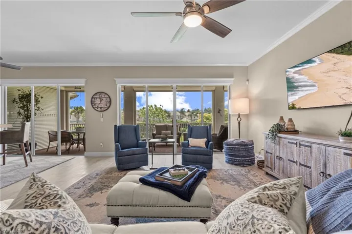 Bright, open living room featuring expansive sliding glass doors framed by transom windows, light wood-finish flooring, a ceiling fan with integrated lighting, and a whitewashed wood-finish built-in media console