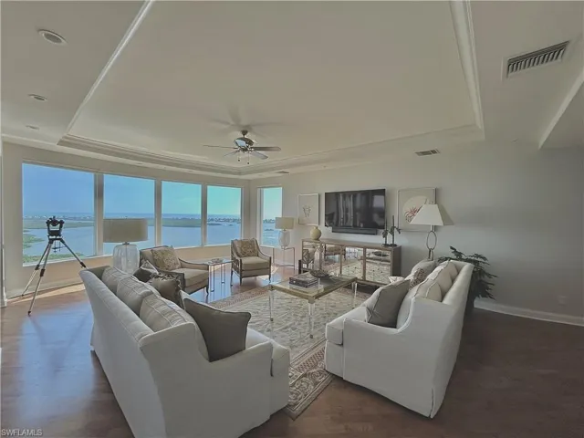 Expansive living space featuring a tray ceiling with recessed lighting, wood-finish flooring, a ceiling fan, and large windows showcasing a water backdrop