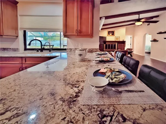 Kitchen with light stone countertops, ceiling fan, lofted ceiling with beams, and a brick fireplace