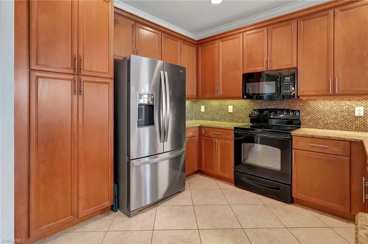 Kitchen with black appliances, brown cabinetry, light stone countertops, backsplash, and ornamental molding