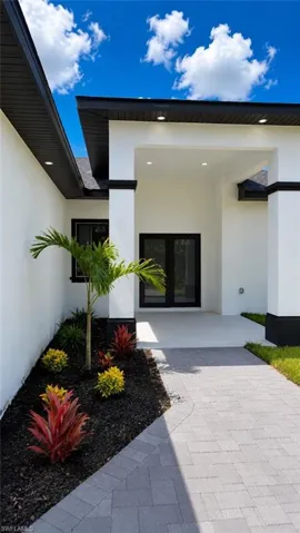 Paved walkway leading to a covered entry with recessed lighting, complemented by landscaping with vibrant foliage and a palm tree