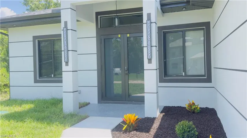 Contemporary exterior featuring a covered entry, dark-framed double glass doors, horizontal paneling, decorative exterior lighting, and a landscaped garden bed