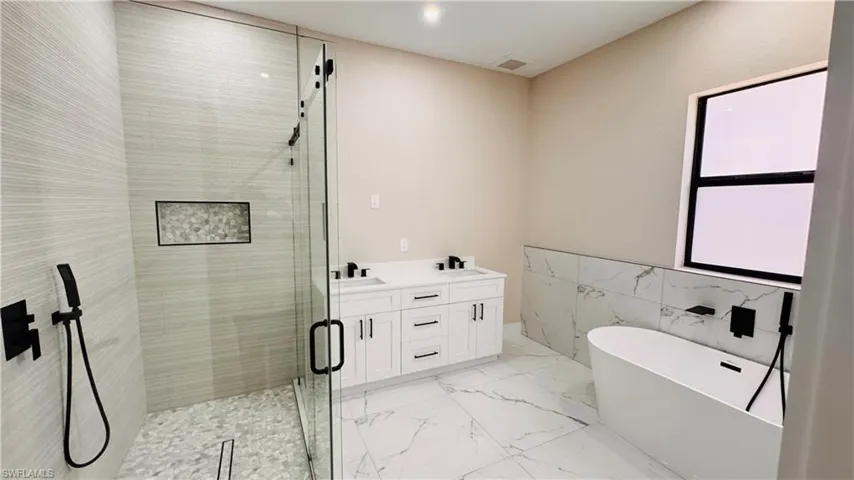 Contemporary bathroom featuring a freestanding tub, a spacious glass-enclosed shower with pebble-style flooring, a double vanity with white cabinetry and dark hardware, large format marble-look floor tiles, and a high window