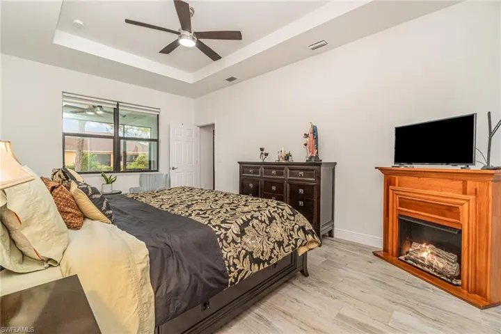 Bedroom with a raised ceiling, light wood-style flooring, a lit fireplace, and ceiling fan