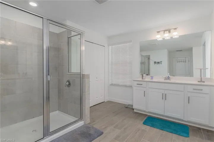 Bathroom featuring a glass-enclosed shower, dual vanity with white cabinetry, large wall-mounted mirror, and wood-finish tile flooring
