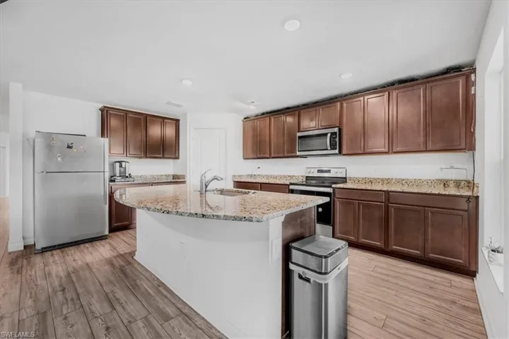 Kitchen featuring stainless steel appliances, light stone counters, dark wood finish cabinetry, light wood finished floors, and a center island with sink