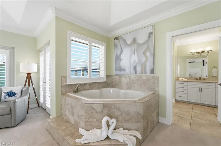 Built-in soaking tub with a tiled surround, plantation shutters, crown molding, and an attached bathroom with a double vanity - Virtually Edited Image