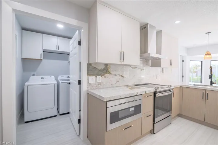 Kitchen with light brown cabinets, pendant lighting, wall chimney exhaust hood, stainless steel electric range oven, and washer and clothes dryer