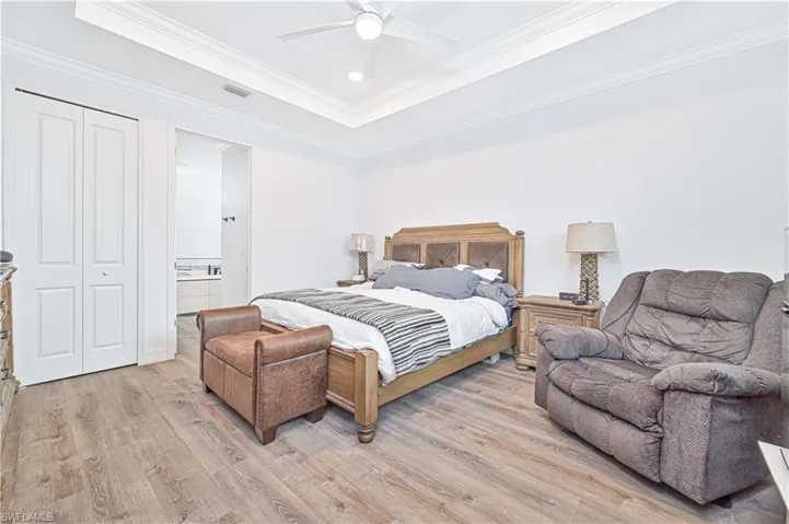 Bedroom featuring light wood finished floors, a raised ceiling, ornamental molding, ensuite bathroom, and ceiling fan