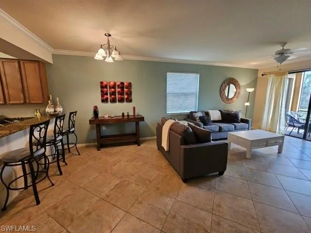 Living room with ceiling fan with notable chandelier, ornamental molding, light tile patterned flooring, and baseboards