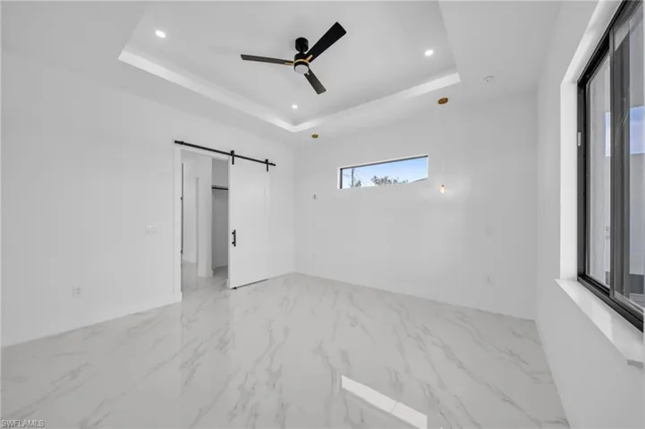 Unfurnished room featuring a barn door, a tray ceiling, recessed lighting, a ceiling fan, and light marble finish floors