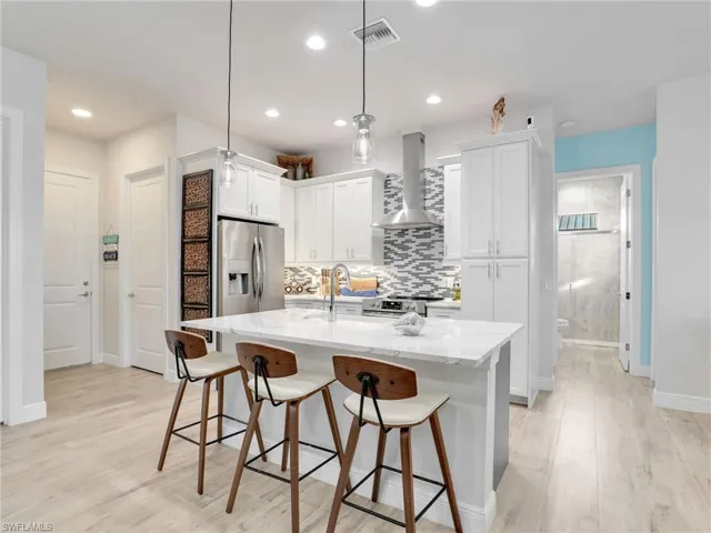 Kitchen featuring hanging light fixtures, wall chimney range hood, an island with sink, stainless steel appliances, and white cabinets