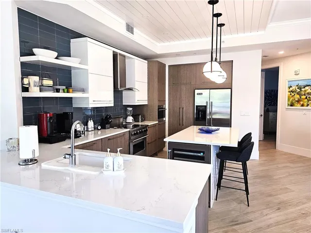 Kitchen with pendant lighting, open shelves, white cabinetry, wall chimney range hood, and a kitchen bar