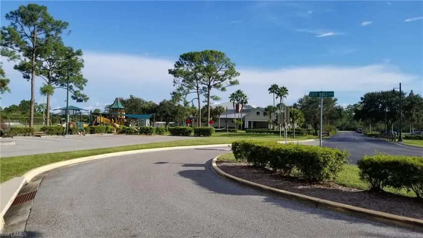 Landscaped community area featuring a children's playground structure with slides and swings, mature trees, paved roadways, and a residential structure with multiple gables and palm trees