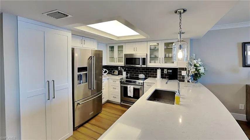 Kitchen featuring white shaker-style cabinetry, stainless steel appliances, a large center island with an undermount sink, a dark subway tile backsplash, and light wood-finish flooring