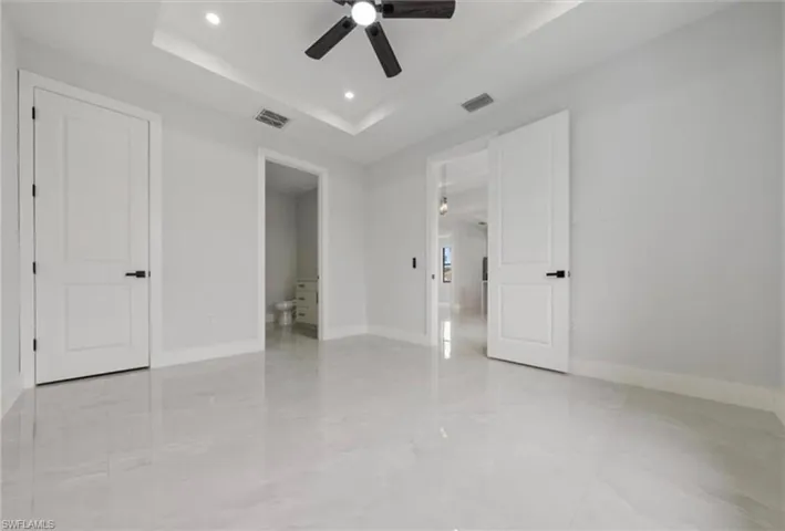 Unfurnished bedroom with a tray ceiling, recessed lighting, a ceiling fan, ensuite bathroom, and light marble finish flooring