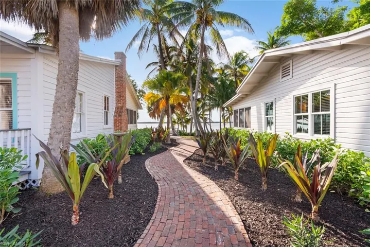 The property features white siding and a brick pathway, surrounded by lush landscaping with palm trees and tropical plants
