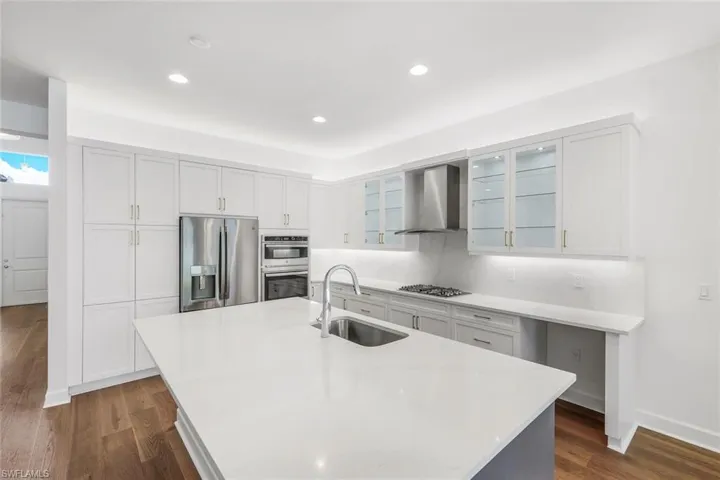 Kitchen with dark wood-type flooring, wall chimney exhaust hood, a kitchen island with sink, sink, and appliances with stainless steel finishes