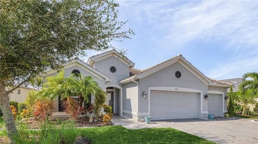 Gray stucco exterior with a tile roof, featuring a paver driveway, an integrated two-car garage, and a lush tropical landscape with mature trees