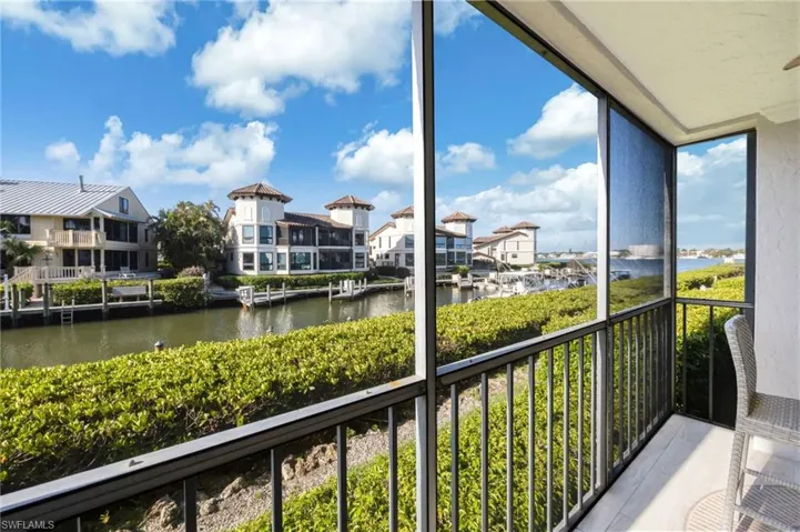 This property features a screened-in balcony with a railing, offering views of a waterway, lush green landscaping, and distant structures