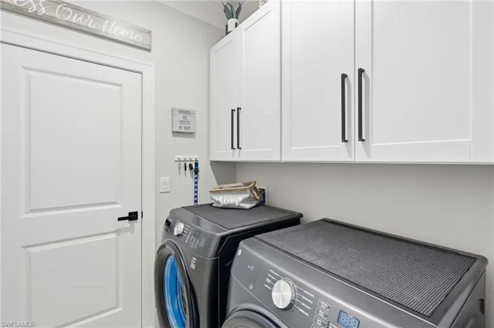 Dedicated laundry room featuring front-load washer and dryer with stainless steel finish, soft-close white cabinetry with brushed metal hardware, quartz countertop work surface, and direct access to the home's interior via solid-panel entry door